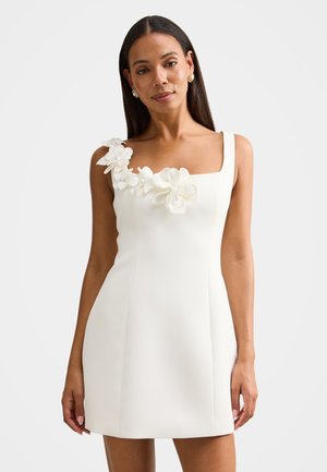 Γυναίκα που φοράει ένα λευκό αμάνικο φόρεμα με τρισδιάστατες ανθικές εφαρμογές στον έναν ώμο και τετράγωνη λαιμόκοψη, στέκεται μπροστά σε μονόχρωμο φόντο.