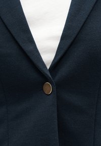 Blazer bleu marine foncé avec un seul bouton métallique sur un t-shirt blanc à col rond, mettant en valeur le revers et le détail du bouton.