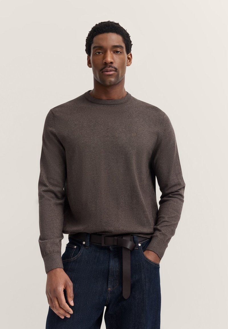 Brauner Langarm-Sweater mit Rundhalsausschnitt aus weichem Material, kombiniert mit blauen Jeans und einem schlichten schwarzen Gürtel. Dezent platziertes Logo auf dem Sweater.