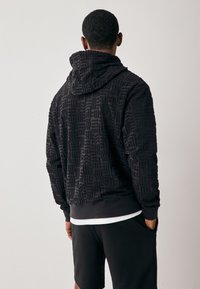 Svart hoodie med texturerat geometriskt mönster, med huva och ribbade muddar, kombinerad med svarta shorts. Minimalistisk design.