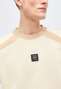 Νεαρός άνδρας που φοράει μπεζ φούτερ με ανοιχτό καφέ επιδιορθώσεις στους ώμους και μαύρο μπλε RB HUGO λογότυπο στο στήθος.