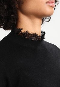 Svart tröja med hög krage som har en delikat svart spetskant. Texturen är mjuk, med en åtsittande passform runt krageområdet.