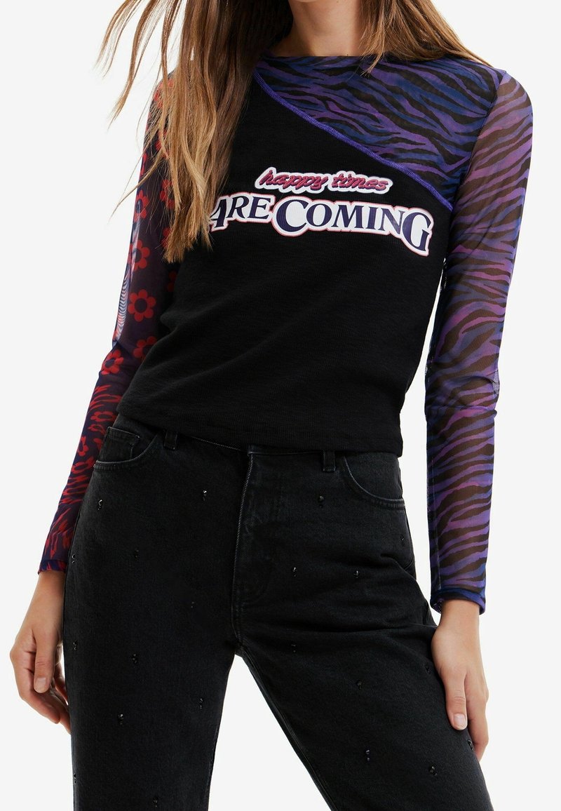 Schwarzes langärmliges Top mit lila tigerstreifigen Netzärmeln, mit dem grafischen Druck "glückliche Zeiten KOMMEN." Kombiniert mit verzierten schwarzen Jeans.