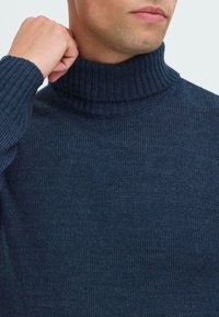 Moški prilagaja ovratnik mornarsko modrega žametanega puloverja s turtleneck, pri čemer je vidno njegovo vrat in spodnji del obraza.