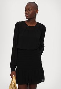 Zwarte jurk met lange, iets bollende mouwen, geglazuurde bodice en gerimpelde zoom. Gedeeltelijk met een gele handtas.