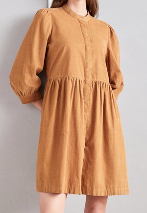 Vrouw draagt een losse, knielange, beige jurk met lange mouwen, een gerimpelde taille en een mandarijnkraag tegen een effen grijze achtergrond.