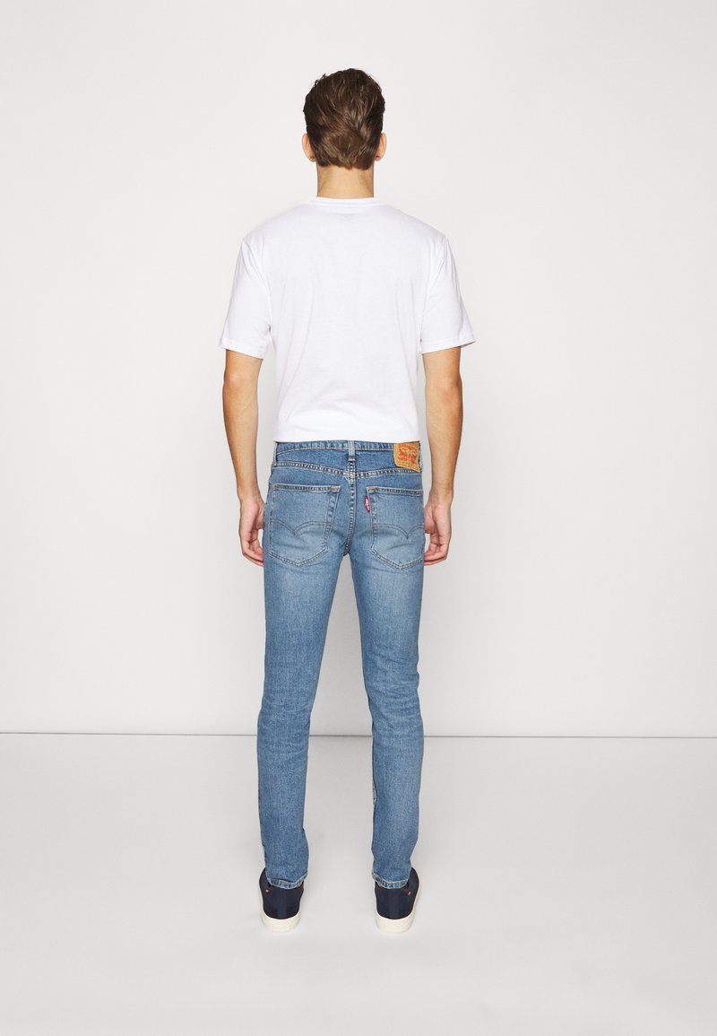 Herren Jeans aus blauem Denim mit Slim Fit, ausgestattet mit fünf Taschen, gesteppten Nähten und einem Leder-Brand-Patch am hinteren Bund. Weißes T-Shirt.