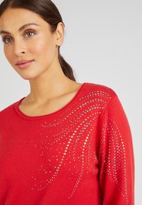 Rode sweater van zachte stof met een ronde halslijn, gekenmerkt door een glanzend patroon van zilveren en gekleurde rhinestones aan één kant.