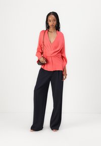 Top avvolgente corallo con maniche lunghe e scollatura a V, abbinato a pantaloni a gamba larga navy a vita alta. Tessuto morbido con vestibilità rilassata.
