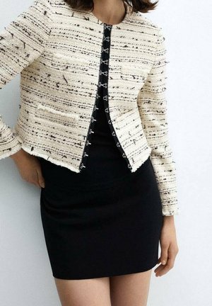 Femme portant une veste courte texturée crème avec des fermetures à crochets noires sur une robe noire ajustée, debout devant un fond blanc.