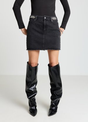 Mini-jupe en denim noir aux bords effilochés, avec passants de ceinture ornés de bijoux. Associée à des bottes noires brillantes montantes jusqu'aux genoux, à bouts pointus.