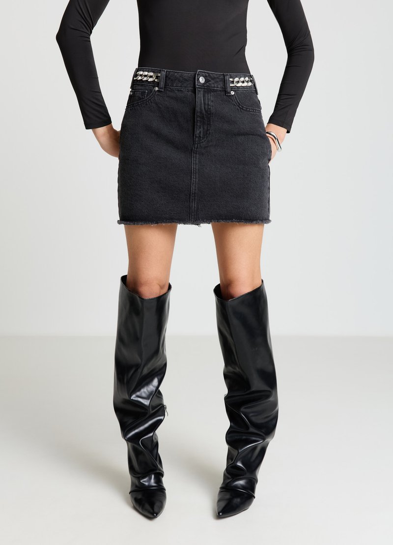 Schwarzer Jeans-Minirock mit ausgefransten Kanten und verzierten Gürtelschlaufen. Kombiniert mit knielangen, glänzenden schwarzen Stiefeln mit spitzen Zehen.