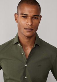 Olijfgroen overhemd met knopen en kraag, voorzien van een subtiel geborduurd logo op de linkerborst en witte knopen. Gladde stoftextuur.