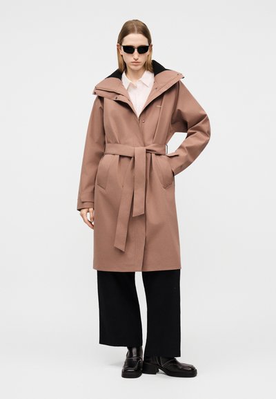 Žena v dlhom mauve páse s opaskom, čiernych nohaviciach s širokými nohavicami, čiernych mokasínach, bielej košeli a čiernych slnečných okuliaroch, stojaca s jednou rukou na bokoch.