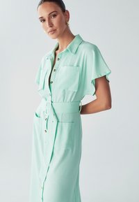 Abito camicia verde menta realizzato in tessuto morbido. Presenta un colletto, chiusura frontale con bottoni, due tasche sul petto e una vita con cintura per definire la silhouette.