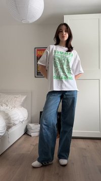 Νεαρή γυναίκα που στέκεται σε υπνοδωμάτιο φορόντας λευκό t-shirt με το σχέδιο "Καλοκαιρινά Όνειρα", μπλε τζιν παντελόνι με φαρδιά μπατζάκια και λευκά αθλητικά παπούτσια.