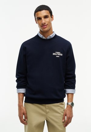 Junger Mann trägt einen marineblauen Pullover mit Rundhalsausschnitt über einem gestreiften Hemd und beiger Hose, steht vor einem einfarbigen weißen Hintergrund.