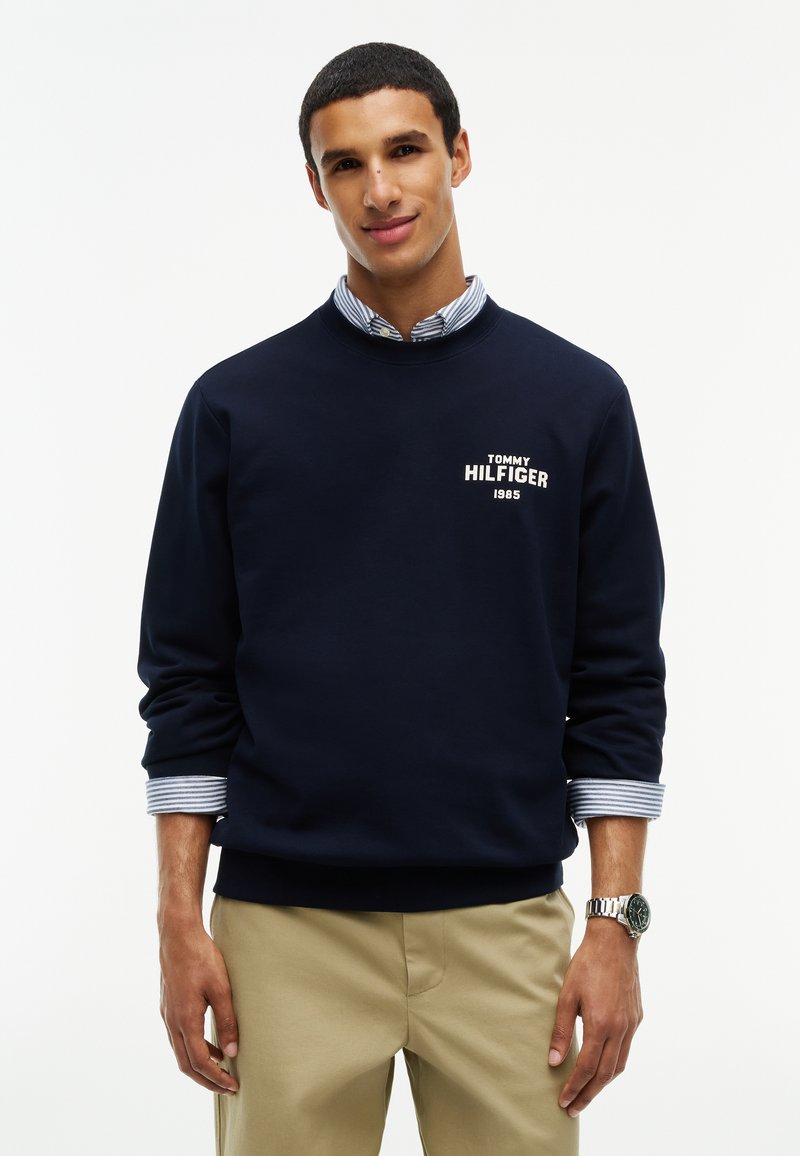 Jeune homme portant un sweat-shirt bleu marine à col rond sur une chemise rayée et un pantalon beige, debout devant un fond blanc uni.