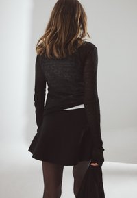 Must must sleeves ja tekstuuriga kangast mustülikond on kombineeritud laieneva musta seeliku ja selja pealt nähtava mudeliga, kes hoiab musta riideeset heledal taustal.