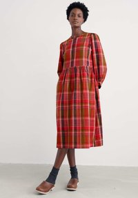 Rot-grünes kariertes Kleid mit langen Ärmeln, durchgehender Knopfleiste, plissiertem Bund und knielanger Länge, kombiniert mit braunen Holzschuhen und grauen Socken.