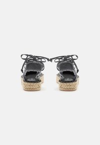 Sandali espadrilla in denim nero con lacci da avvolgere alla caviglia e suola platform intrecciata in juta, visti da dietro su sfondo bianco.