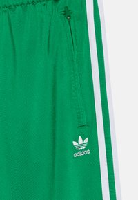 Πράσινες αθλητικές βερμ�ούδες από λείο ύφασμα, με τσέπη με φερμουάρ, λευκές ρίγες στο πλάι και ένα μικρό λευκό λογότυπο της Adidas στο κάτω αριστερό μέρος.