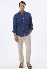 Camisa azul de manga larga con cuello de botón y botones blancos, combinada con pantalones beige claros y sandalias marrones.