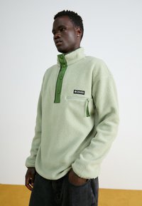 Lys grønn fleece-pullover med grønn knappelukking krage, glidelåslomme på brystet og en svart logo-patch på venstre side.