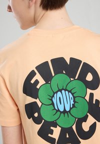 Persikka puuvillapaita, jossa on suuri musta grafiikka, jossa on teksti "LÖYDÄ RAUHASI", ympäröitynä vihreällä kukkakuviolla.