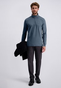 Uomo con una maglia blu a maniche lunghe con zip, pantaloni grigi, scarpe nere, che tiene un cappotto nero nella mano sinistra, in piedi su uno sfondo neutro.