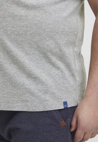 Tricou din bumbac gri, cu guler rotund și mâneci scurte. Prezintă o etichetă mică de marcă albastră în partea stângă jos.