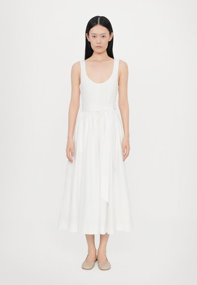 Ärmelloses weißes Leinenkleid mit taillierter Oberteile, fließendem Midiskirt und einem selbstgebundenen Gürtel in der Taille. Fußbekleidung in neutralen Farben.