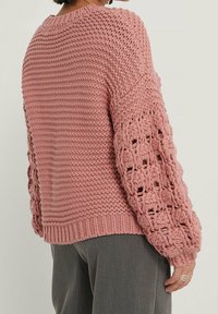 Rosa stickad tröja med texturerad kropp, puffärmar med luftiga mönster och ribbad nederkant. Tyget verkar tjockt och mysigt.