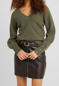Kobieta nosi oliwkowy sweter dziergany z zapięciem na guziki przy mankietach oraz czarną skórzaną mini spódnicę z zamkiem błyskawicznym z przodu i sześciokątną klamrą pasa.
