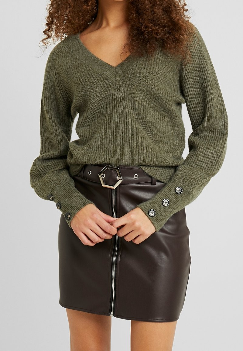 Kobieta nosi oliwkowy sweter dziergany z zapięciem na guziki przy mankietach oraz czarną skórzaną mini spódnicę z zamkiem błyskawicznym z przodu i sześciokątną klamrą pasa.