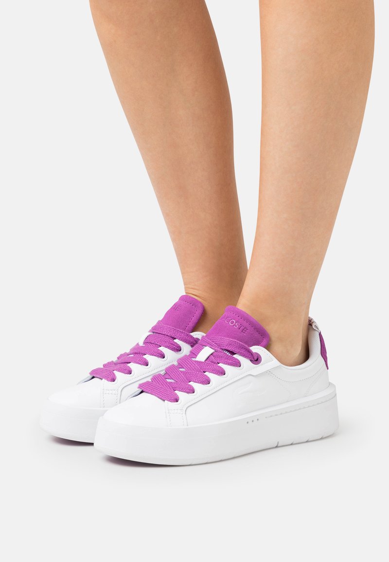 Weiße Sneaker mit lila Schnürsenkeln und Akzenten, ausgestattet mit einem glatten Lederoberteil, flacher Sohle und minimalistischen Design.