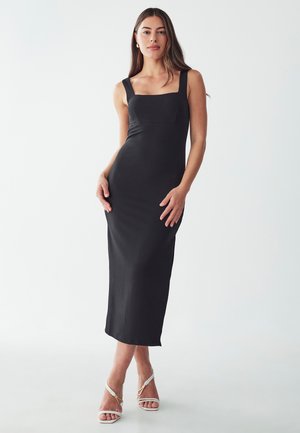 Black, fitted midi dress with square neckline and wide straps. Smooth texture, simple design, paired with white strappy heels.