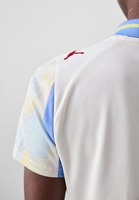 Maillot blanc avec une texture en mesh, présentant un motif abstrait bleu pastel et jaune sur les manches. Logo rouge sur l'épaule arrière.