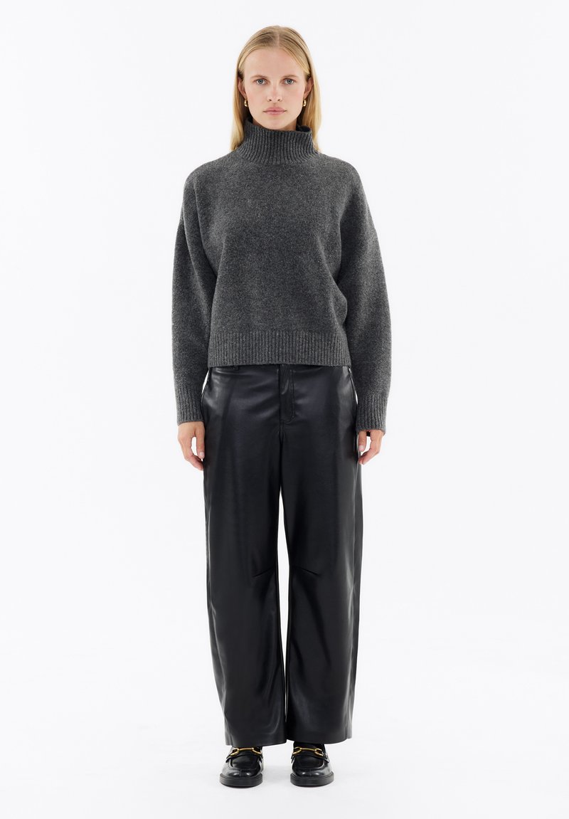 Dunkelgrauer Strickpullover mit hohem Kragen, kombiniert mit schwarzer, lederner Schlaghose. Das Outfit zeichnet sich durch einen lässigen Schnitt und minimale Details aus.
