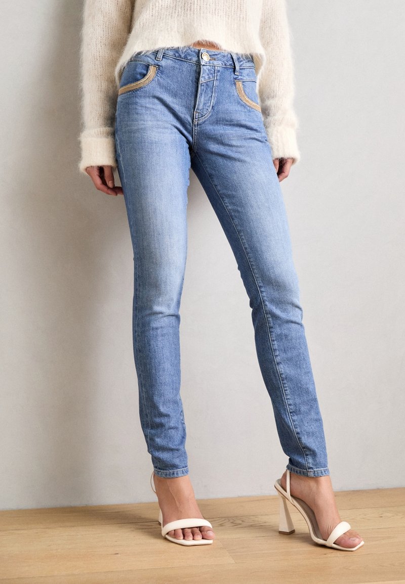 Personne portant un jean skinny bleu clair et des sandales blanches à talons hauts, debout sur un sol en bois devant un mur gris uni.