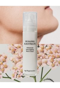 Biała cylindryczna butelka z pompką, oznaczona "Susanne Kaufmann Advanced Anti-Ageing Rich Cream", otoczona różowymi kwiatami i skórą.