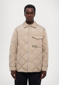 Giubbotto imbottito beige con motivi a rombi, chiusura con bottoni, una tasca sul petto e una discreta etichetta con logo sul lato. Tessuto strutturato, vestibilità rilassata.