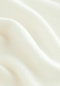Teksturowana biała tkanina z miękkim, tkanym wzorem. Powierzchnia wydaje się gładka z niewielkimi różnicami w grubości, co tworzy wizualną głębię.