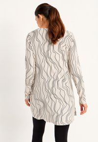 Vrouw met bruin haar in een paardenstaart, gekleed in een lange beige tuniek met zwarte golvende lijnen, gecombineerd met zwarte legging, met haar rug naar ons toe gekeerd.