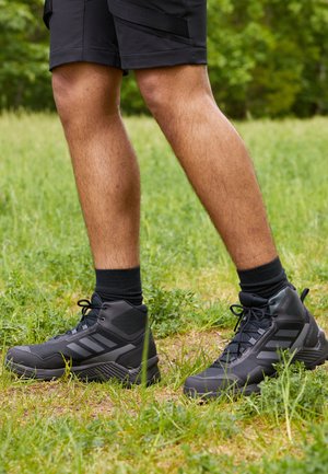Zwarte hoge sportieve schoenen met een textuur, een drie-strepen ontwerp en zwarte veters, gecombineerd met zwarte korte sokken en shorts.