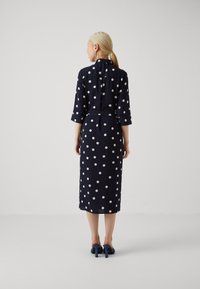 Rochie bleumarin cu buline, cu mâneci trei sferturi, talie încinsă și tiv până la genunchi. Textură de stofă netedă. Asortată cu pantofi cu toc bleu.
