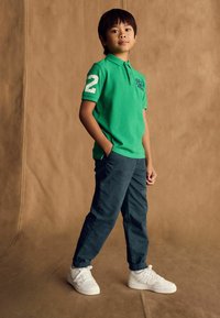 Groene polo met een 2 op de mouw, gecombineerd met donkere broeken en witte sneakers. Soepele textuur en casual pasvorm.