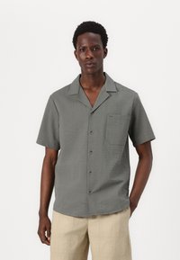 Camisa de manga corta gris con un patrón texturizado, cierre de botones en el frente y un bolsillo en el pecho. Combinada con pantalones texturizados de color beige claro.