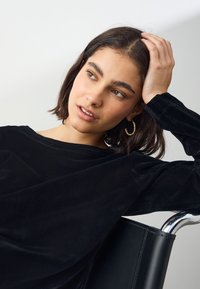 Haut à manches longues en velours noir avec une coupe décontractée, présentant un large décolleté et une texture lisse. Porté avec de grandes boucles d'oreilles en or.