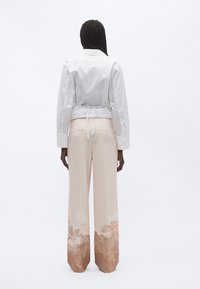 Balta īsa jaka ar jostu un garām piedurknēm, kopā ar plašām krēmkrāsas biksēm ar rozā ziedu rakstu apakšmalā.